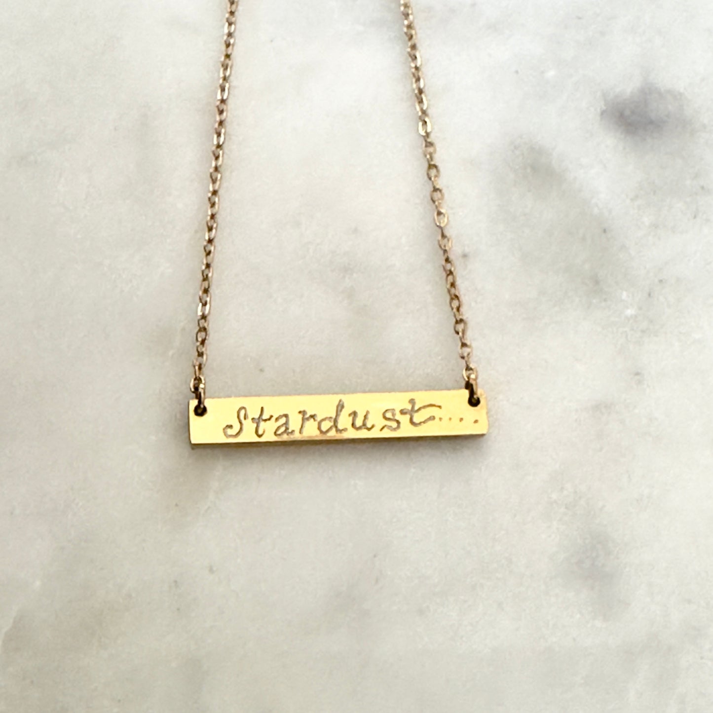 Gold bar necklace with 'Stardust' script on a marble surface