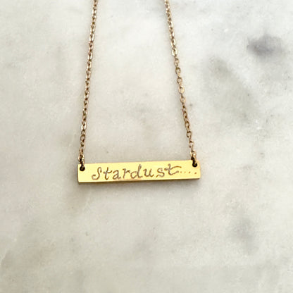 Gold bar necklace with 'Stardust' script on a marble surface